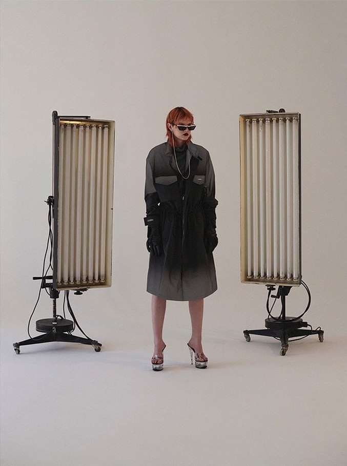 A stylish adult female with reddish hair and sunglasses poses in a modern ombre coat, platform heels, and vintage light stands in a studio setting.