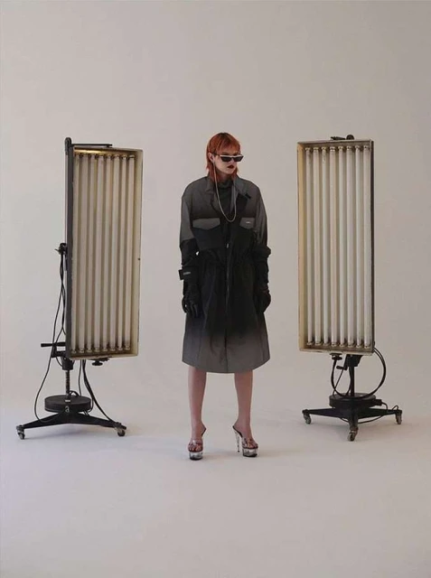 A stylish adult female with reddish hair and sunglasses poses in a modern ombre coat, platform heels, and vintage light stands in a studio setting.