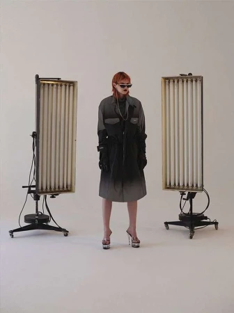 A stylish adult female with reddish hair and sunglasses poses in a modern ombre coat, platform heels, and vintage light stands in a studio setting.