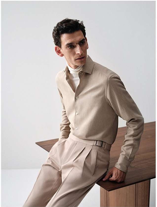 Studio shot of a male model wearing a beige button-down shirt with long sleeves, beige pants, and a white turtleneck. He's leaning on a table.