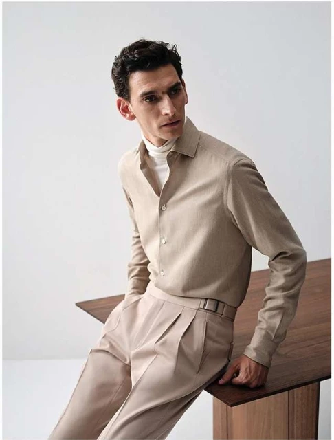 Studio shot of a male model wearing a beige button-down shirt with long sleeves, beige pants, and a white turtleneck. He's leaning on a table.