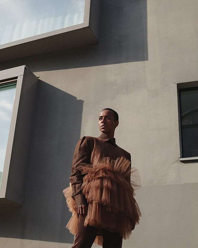 Model with short hair wearing a brown shirt and layered tulle skirt outdoors against a grey modern building wall.
