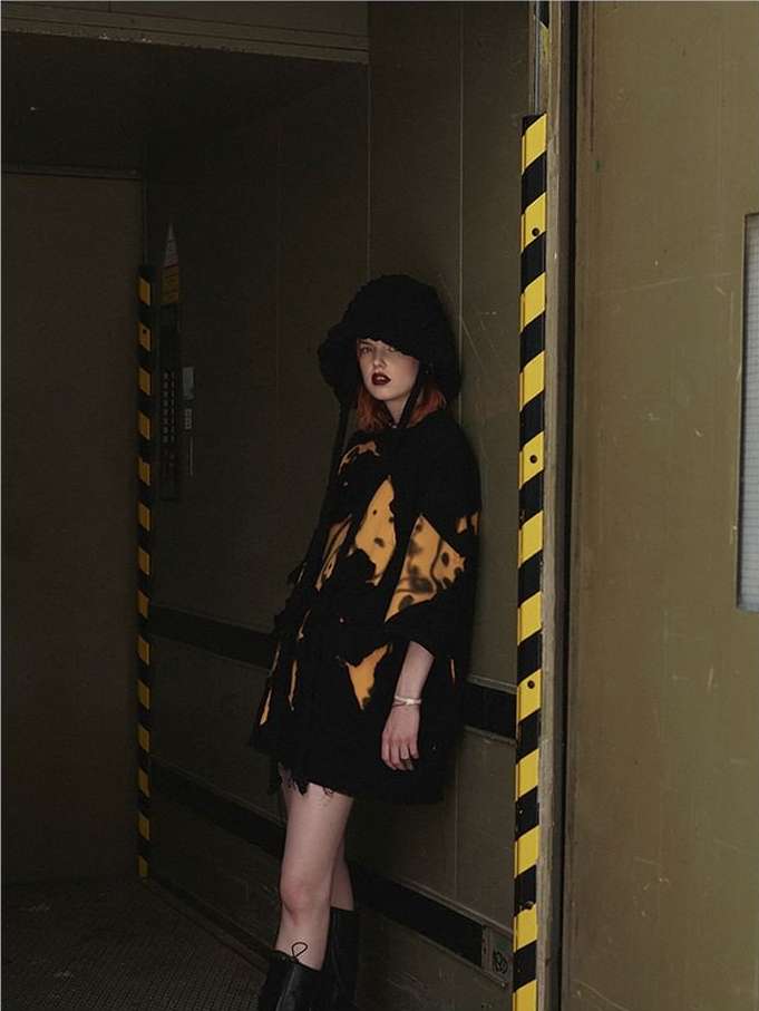 A trendy woman in a bucket hat and patterned jacket poses near a metal door with hazard stripes in an industrial indoor setting.