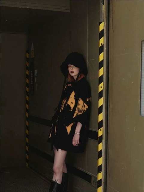 A trendy woman in a bucket hat and patterned jacket poses near a metal door with hazard stripes in an industrial indoor setting.