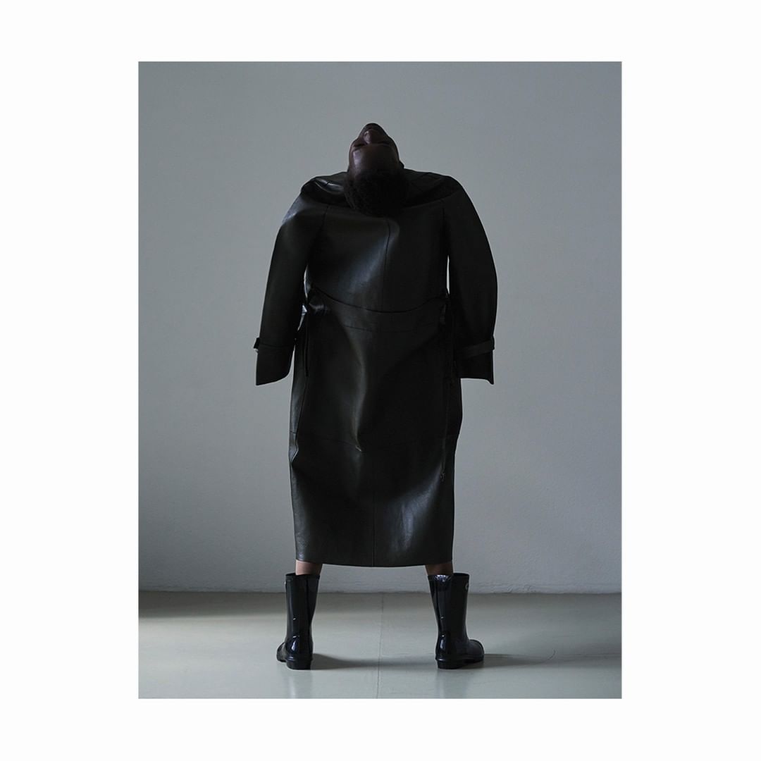 A figure in a long, dark leather coat and boots stands against a grey wall, facing away from the viewer with their head tilted back.