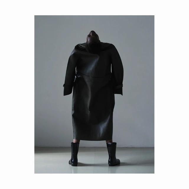 A figure in a long, dark leather coat and boots stands against a grey wall, facing away from the viewer with their head tilted back.