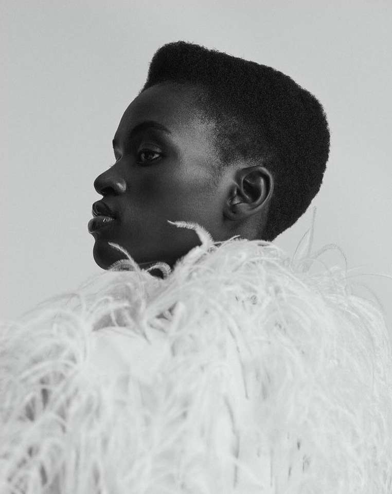 Monochromatic close-up portrait of a Black female model in a profile pose wearing an extravagant white feathered garment against white backdrop.