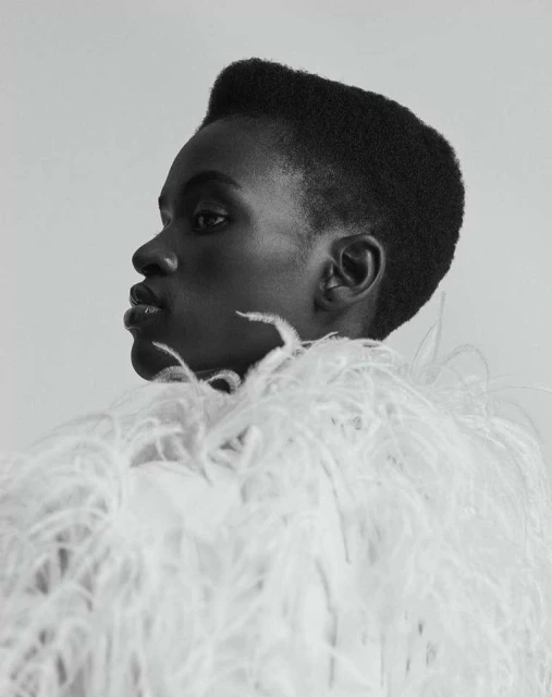 Monochromatic close-up portrait of a Black female model in a profile pose wearing an extravagant white feathered garment against white backdrop.