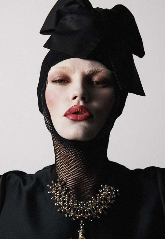 Close-up of a fashion model wearing a black head covering with a large bow, and a beaded gold necklace, against a light background.