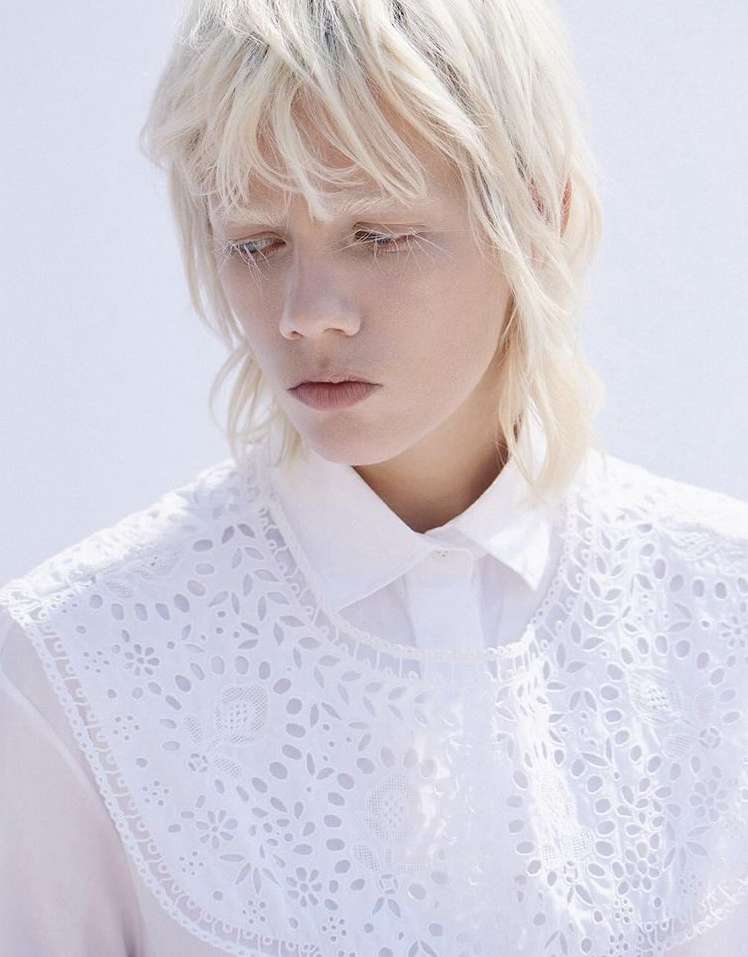 Close-up portrait of an adult female with short blonde hair wearing a white eyelet-trimmed shirt against a light blue background.