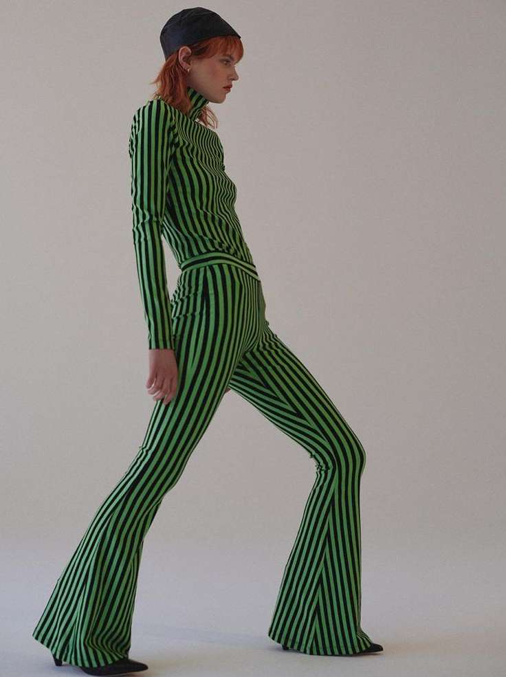 Side profile of a stylish adult woman in a green and black striped two-piece outfit and black cap walking in a brightly lit photo studio.