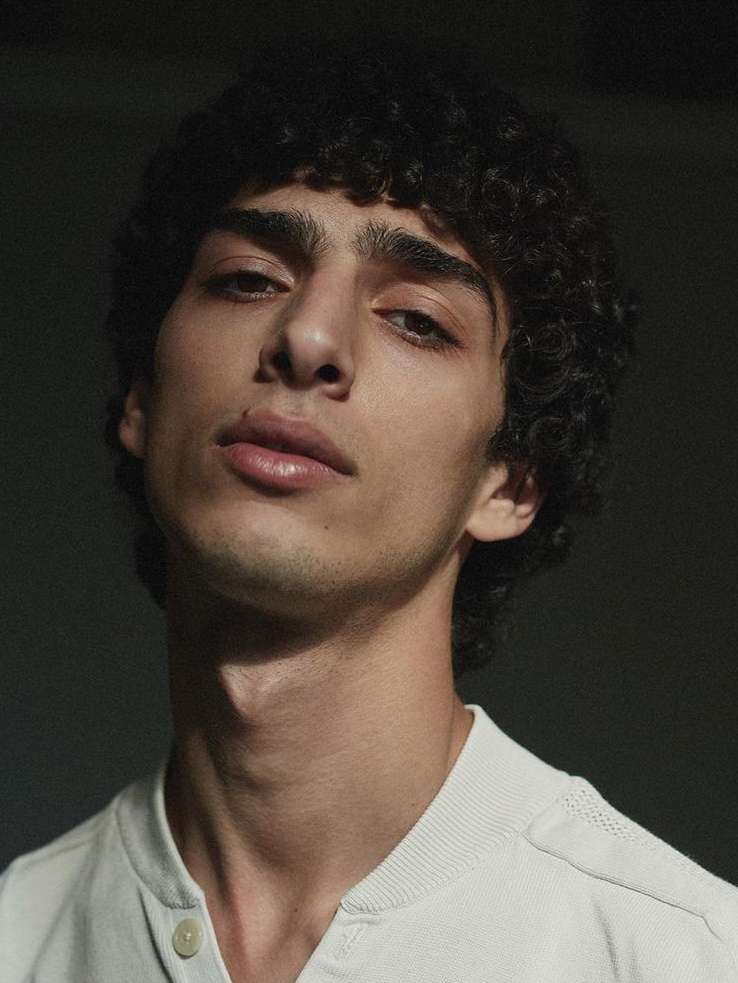 Portrait of a dark-haired young adult male with curly hair looking at the camera, wearing a white shirt, against a dark background.