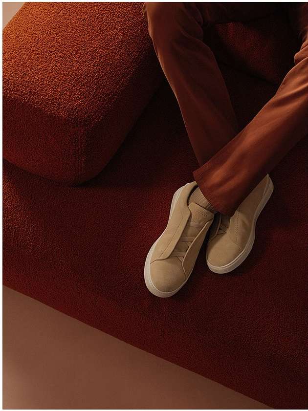 Close-up of cream sneakers and brown trousers on a textured rust-coloured upholstered sofa by Ansgar Sollmann
