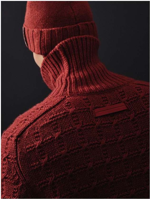Close-up studio shot of a person from the back wearing a red textured turtleneck sweater and a matching beanie hat against a dark background.