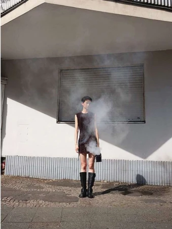 Model standing outdoors near a building exterior wearing a dark dress and black boots amidst drifting smoke by Ansgar Sollmann