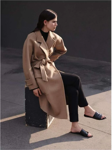 Elegant woman in a tan trench coat sitting on a concrete block, showcasing a minimalist fashion style against a grey background.