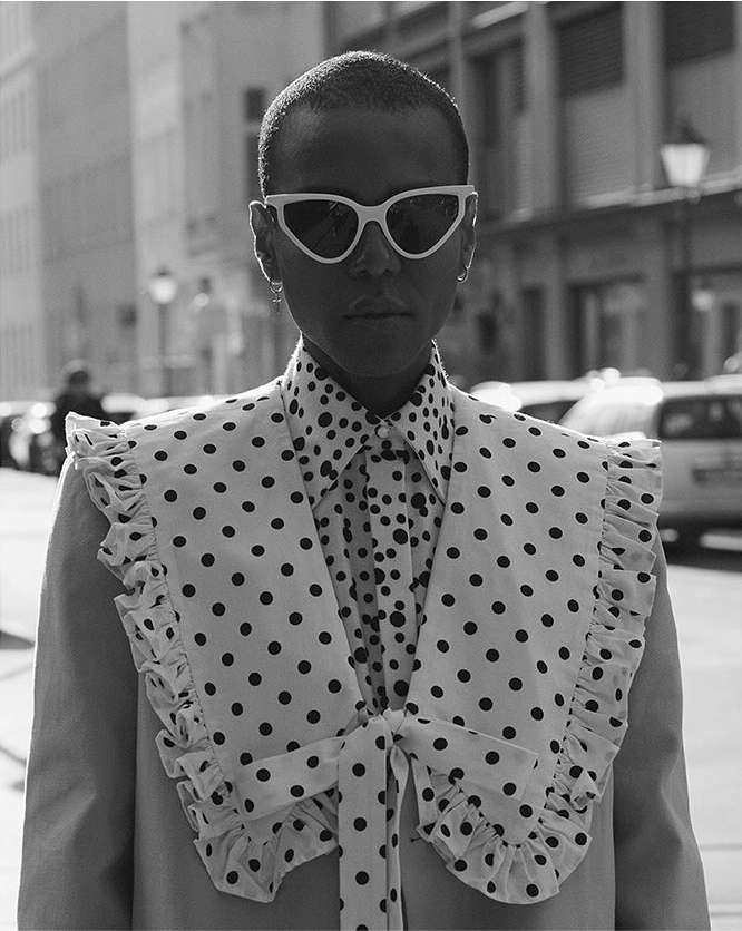 Black and white portrait of a person with sunglasses, wearing a polka dot shirt and ruffled collar standing in an urban setting.