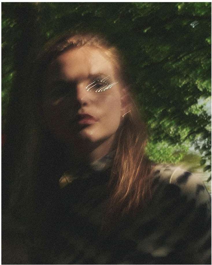 A young adult woman with light brown hair looks at the camera, outdoors. The foreground and shadows surround the subject.