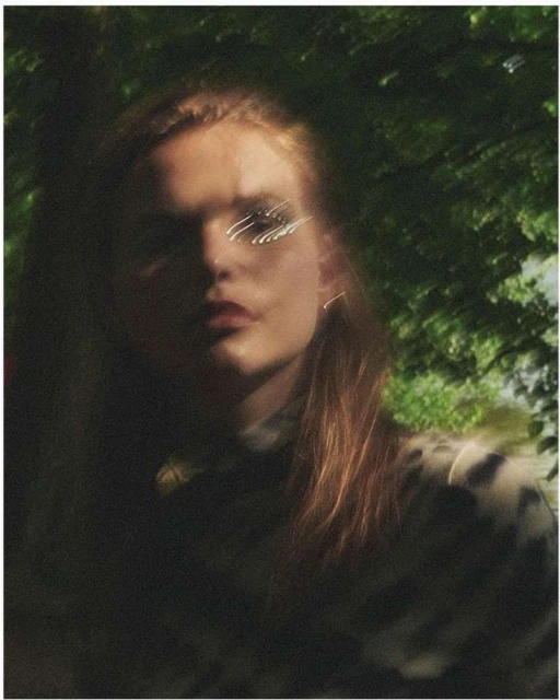A young adult woman with light brown hair looks at the camera, outdoors. The foreground and shadows surround the subject.