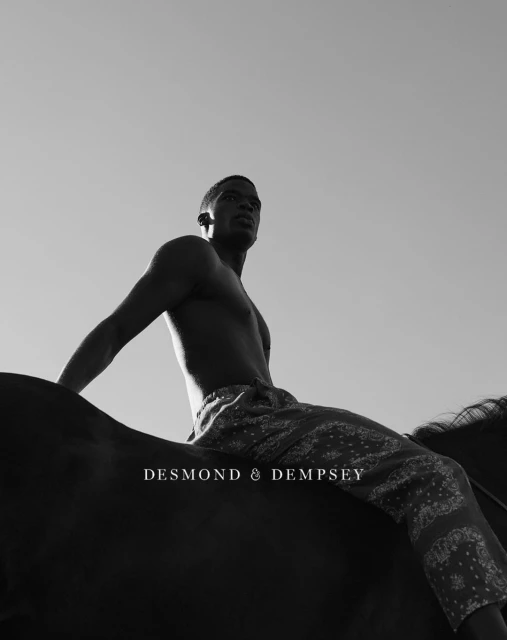 Low angle, black and white shot of a shirtless adult male in paisley patterned pants on horseback beneath a clear sky.