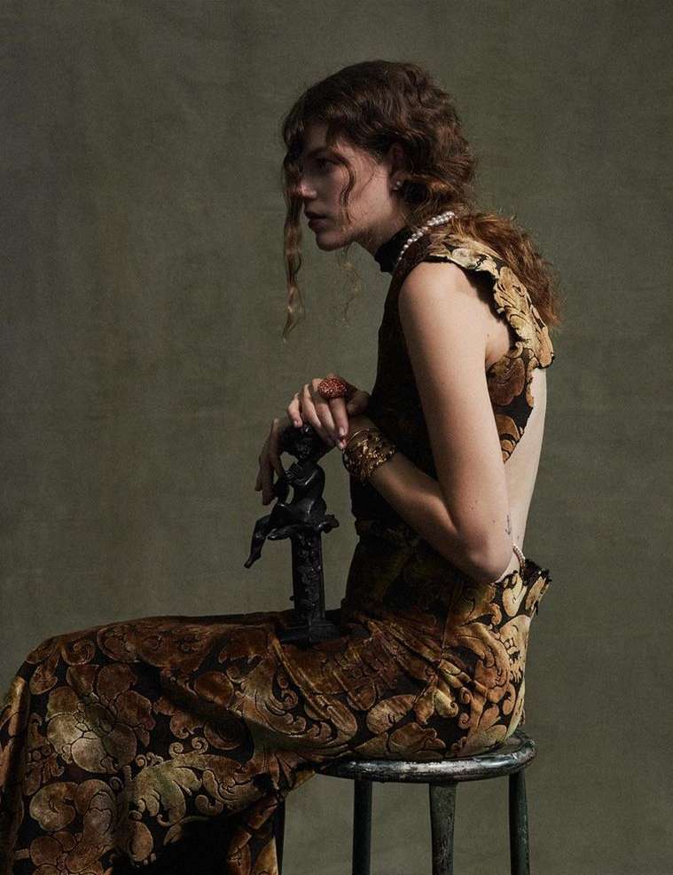 Elegant female model in patterned dress sits holding sculpture in studio, facing left with soft focus and muted tones for fashion editorial.