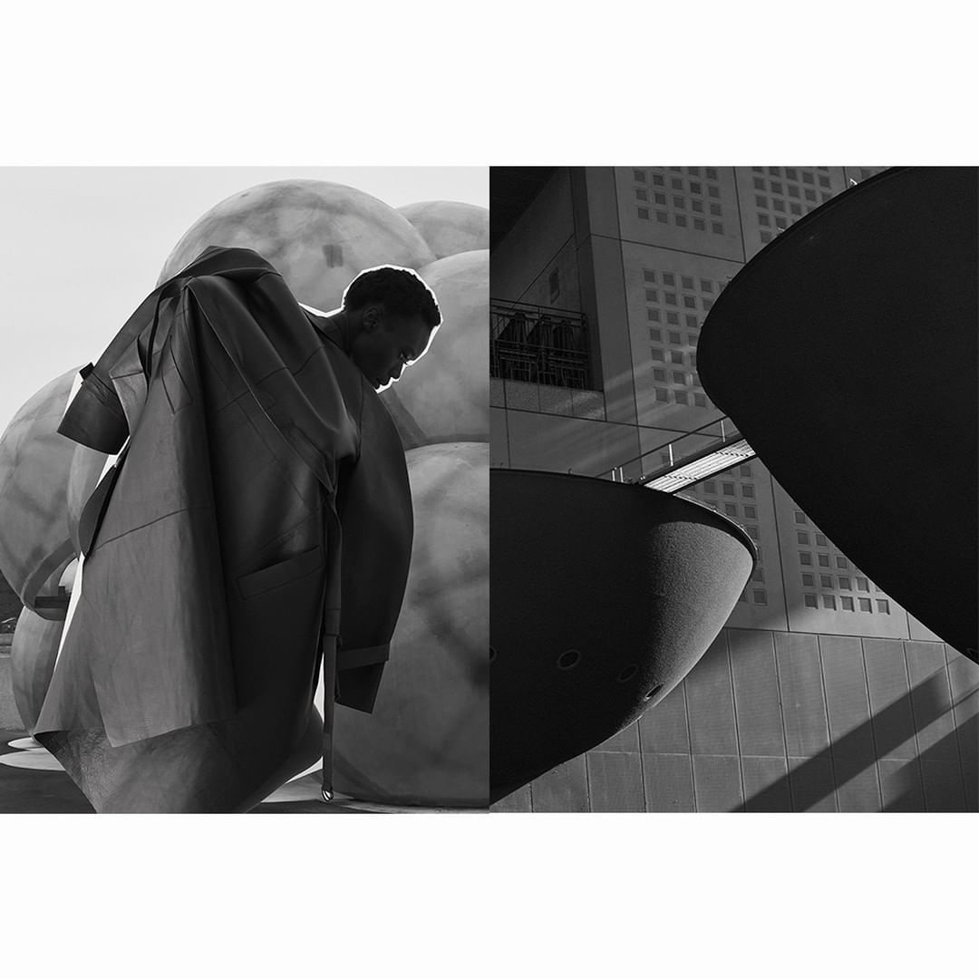 Monochrome fashion shot of a Black man in a coat beside large spheres and modern architecture, captured with a wide shot.