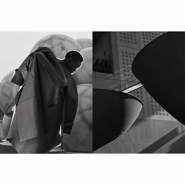 Monochrome fashion shot of a Black man in a coat beside large spheres and modern architecture, captured with a wide shot.