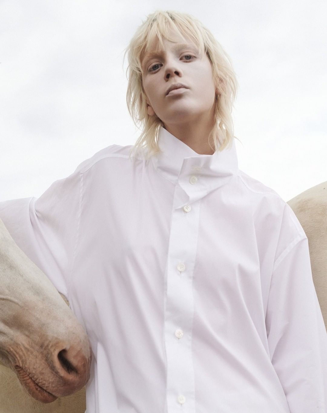 Portrait of a blonde model wearing a white buttoned shirt standing beside a horse against an overcast sky