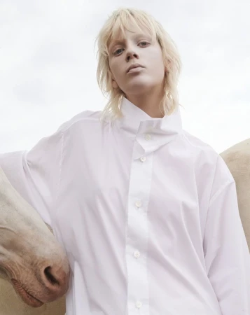 Portrait of a blonde model wearing a white buttoned shirt standing beside a horse against an overcast sky