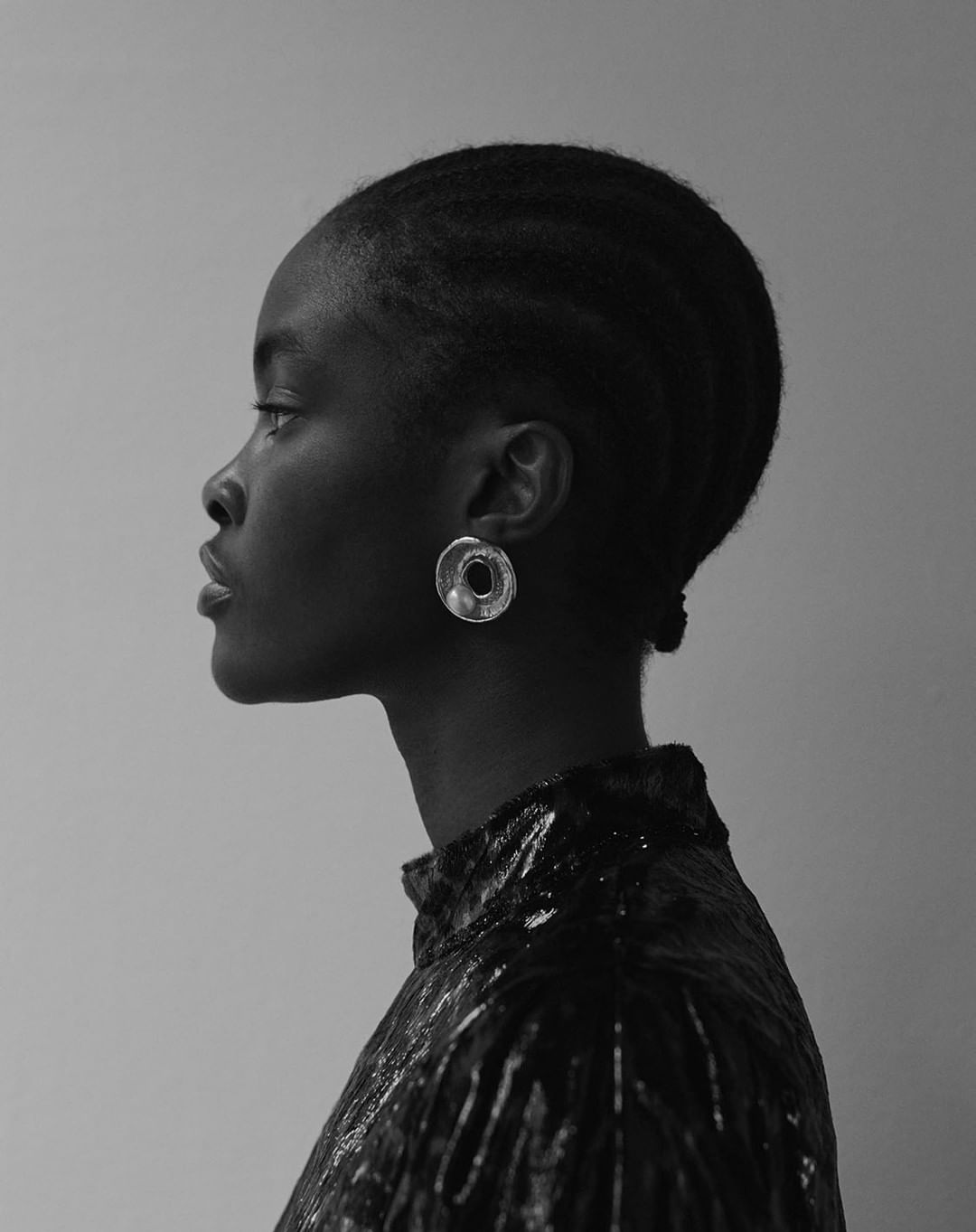 Close-up profile of an adult female with dark skin and braided hair, wearing round metallic earrings and a high-necked black top.