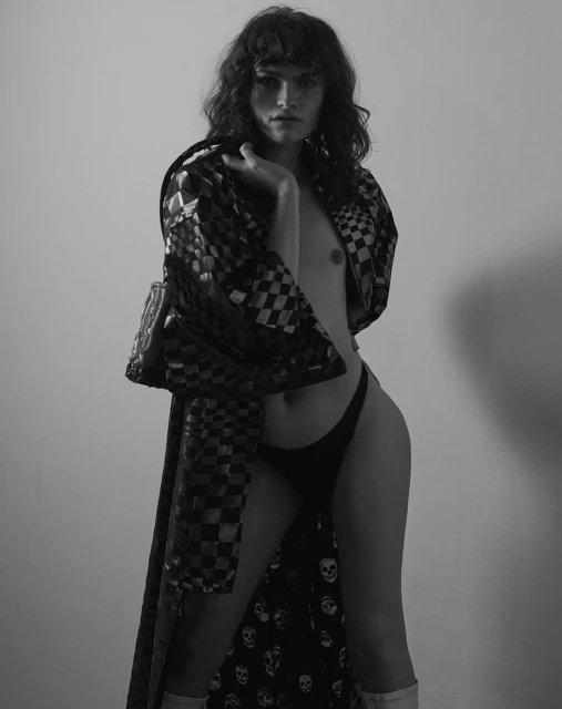 Monochrome portrait of a slim adult posing in a patterned robe and minimal bottoms in a photo studio setting with soft lighting.