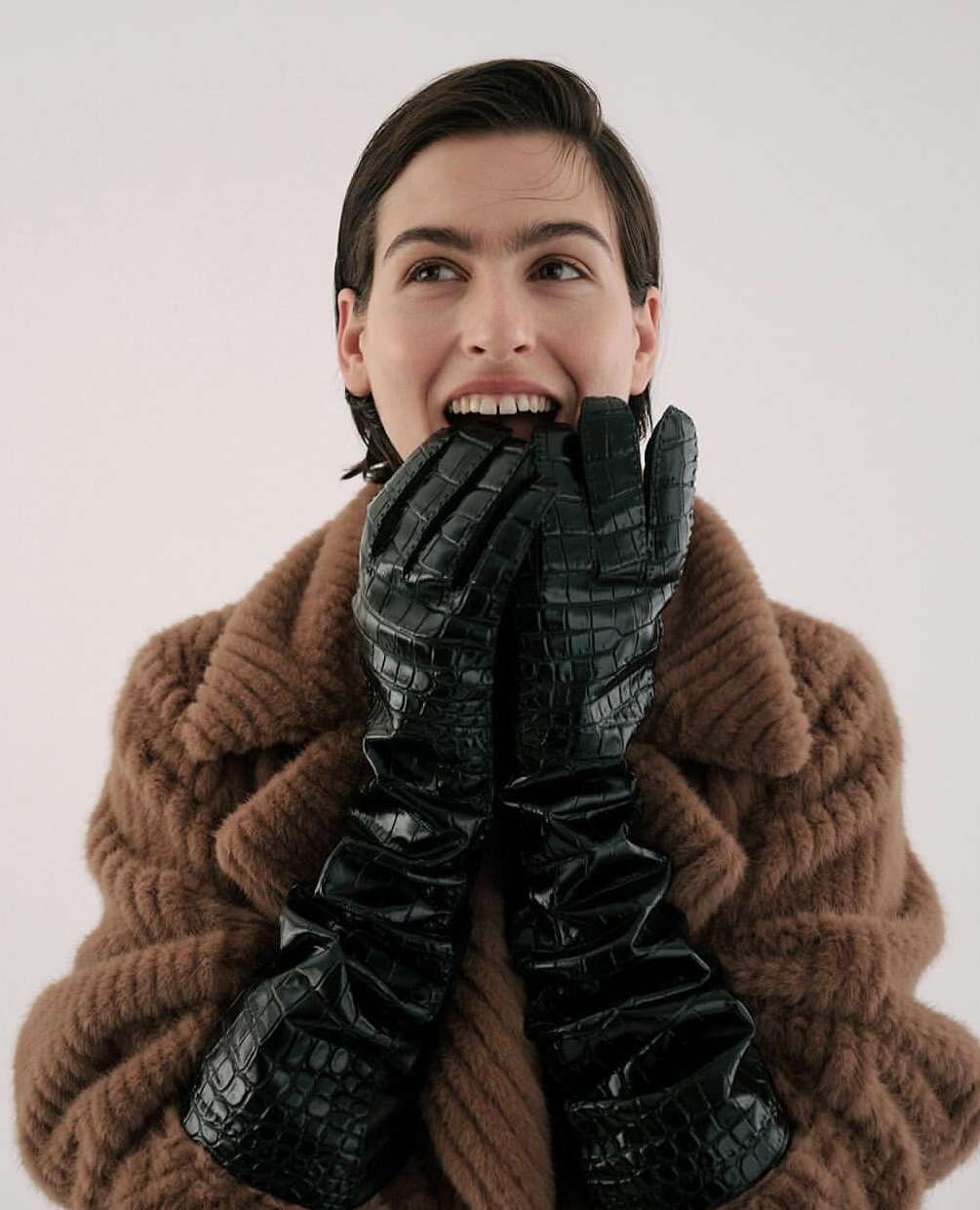 A woman with short brown hair, wearing a brown coat and black crocodile-texture gloves, has her hands near her mouth in an indoor studio setting.