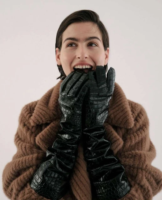 A woman with short brown hair, wearing a brown coat and black crocodile-texture gloves, has her hands near her mouth in an indoor studio setting.