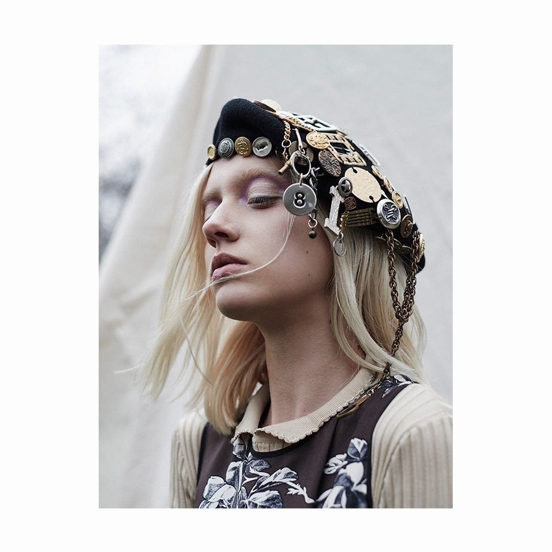 Portrait of woman in embellished beret with eyes closed. She wears a floral dress and neutral sweater in an outdoor environment.