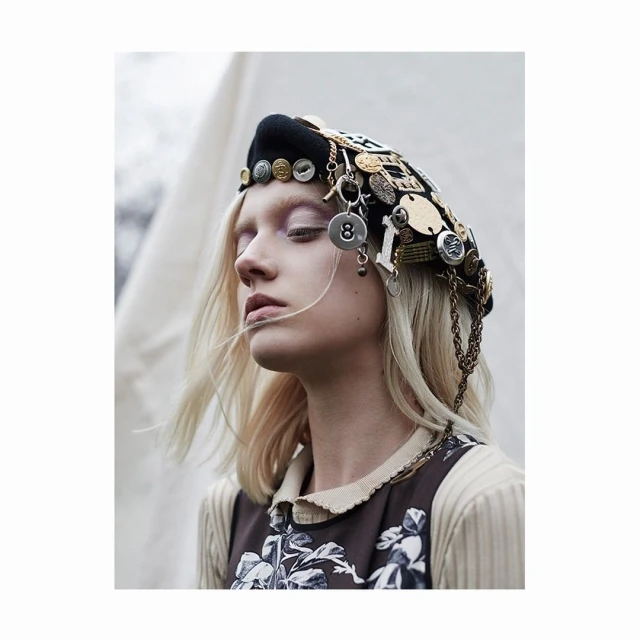 Portrait of woman in embellished beret with eyes closed. She wears a floral dress and neutral sweater in an outdoor environment.