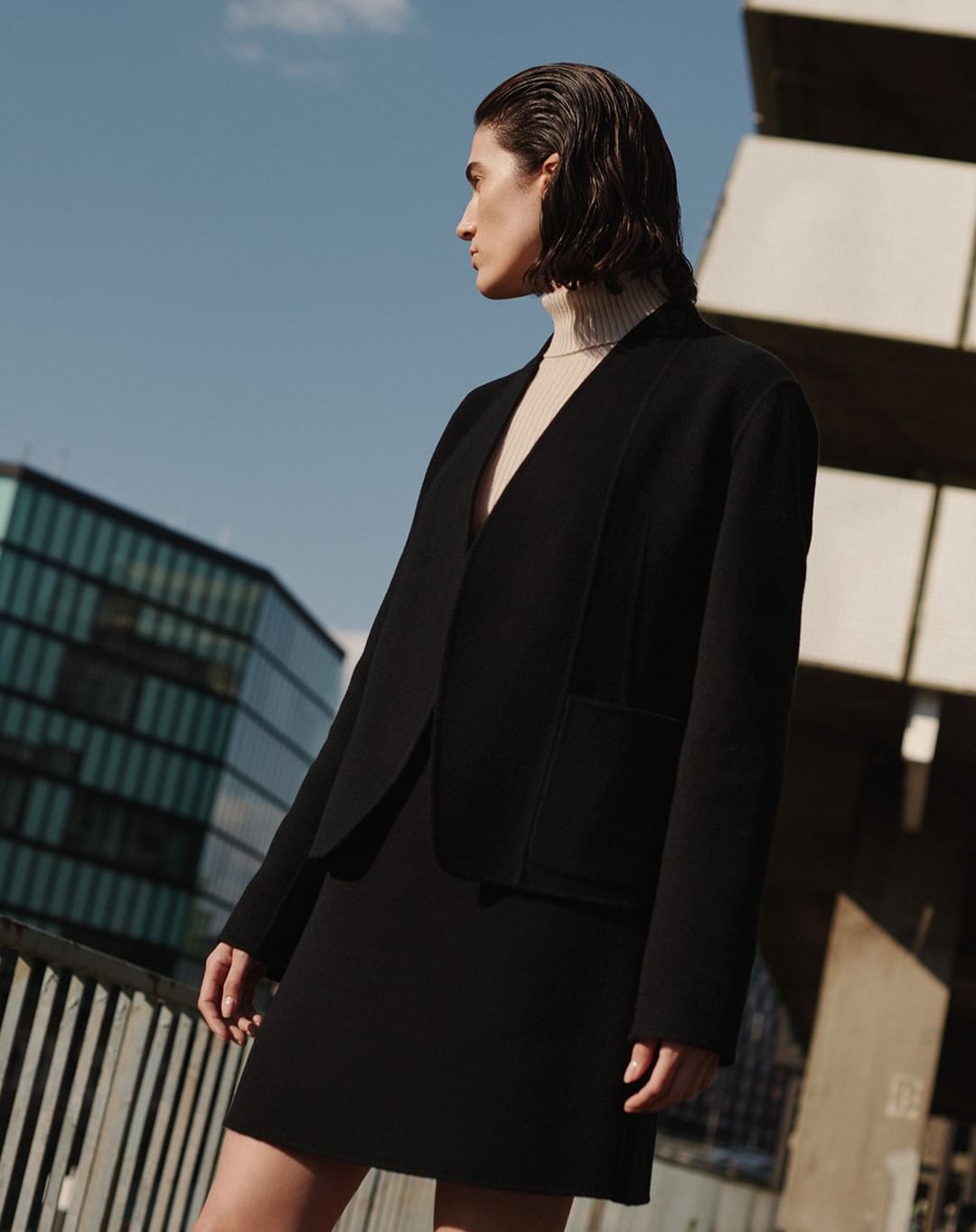 Model with slicked-back dark hair wearing a black blazer and matching skirt outdoors against a clear blue sky.