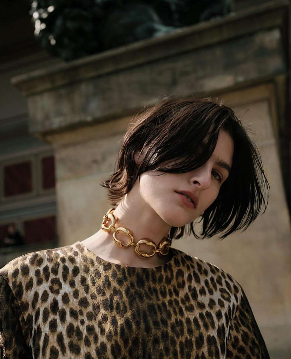 Fashion shot of a model in a leopard print top and gold necklace against a stone background, captured in natural light.