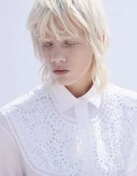 Portrait of a blonde model with bleached eyelashes wearing a white eyelet embroidered shirt against a bright background