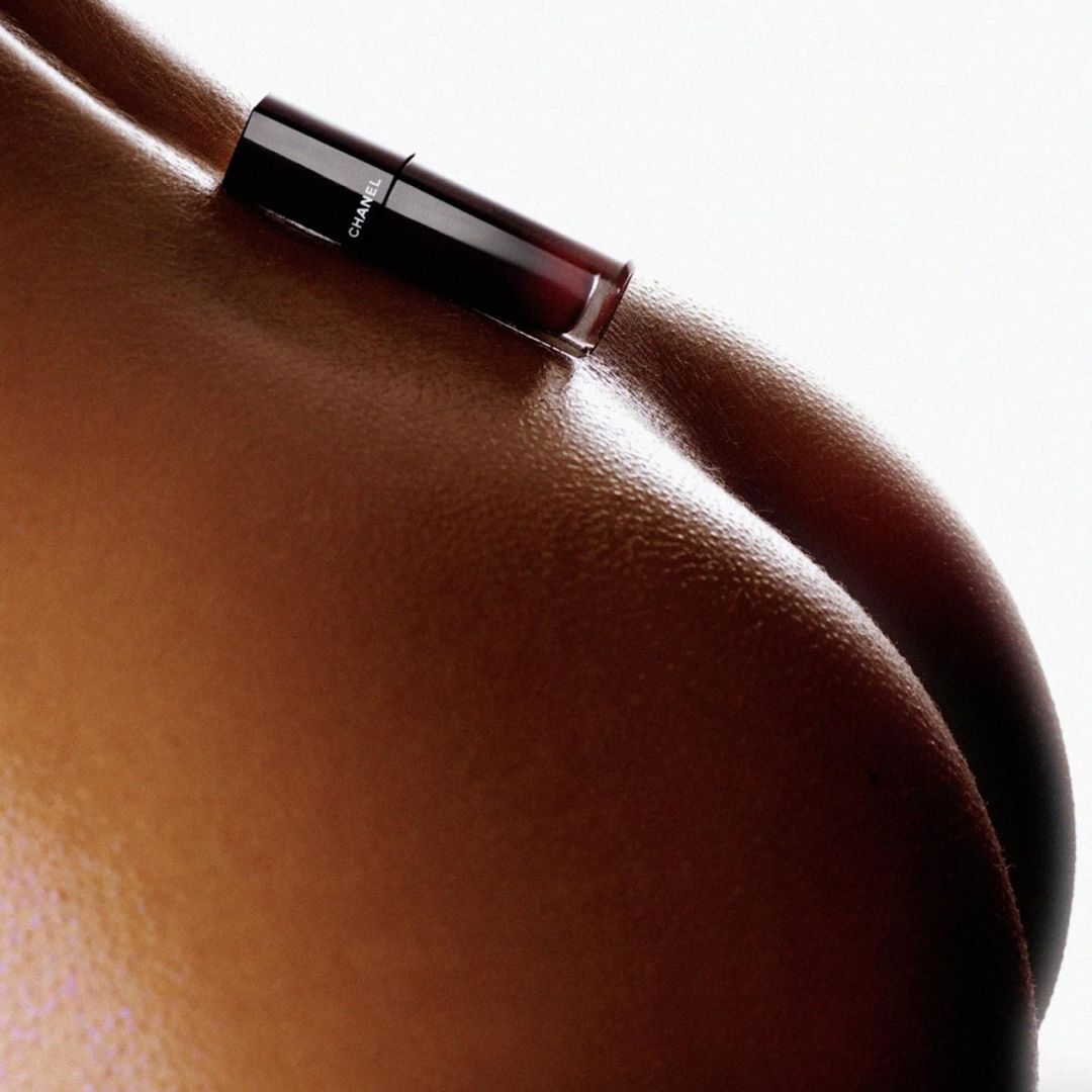 Close-up studio shot of tanned skin with a black Chanel lip gloss product, emphasizing beauty, fashion, and cosmetic aesthetics.