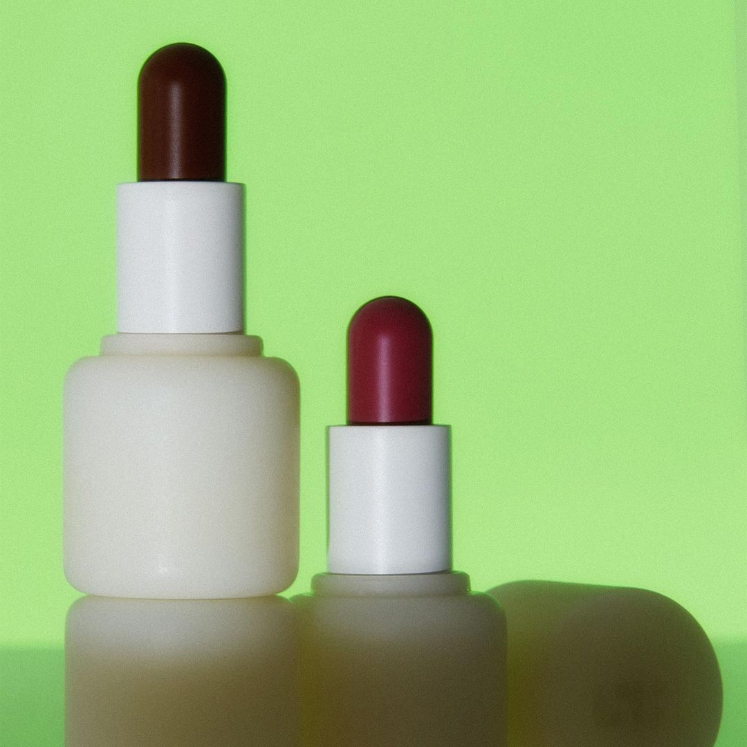 Two cosmetic bottles, one larger, with brown caps, sit on a reflective surface against a bright green background, product photography.