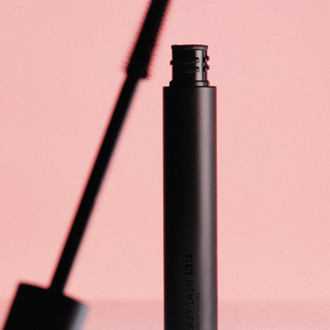 A black mascara tube with a visible brush sits against a soft pink background in a beauty product shot showcasing cosmetic design.