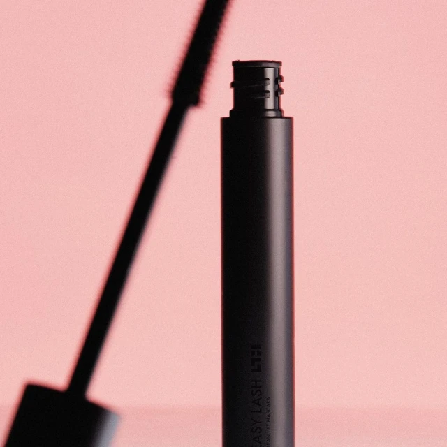 A black mascara tube with a visible brush sits against a soft pink background in a beauty product shot showcasing cosmetic design.