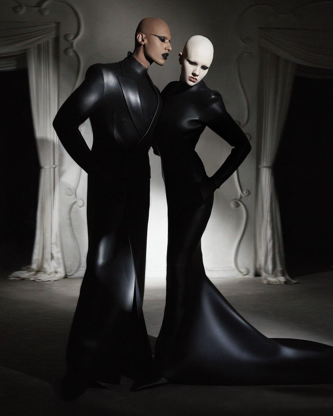 Two bald fashion models in matching black gowns pose in a studio with ornate white molding and soft lighting, evoking avant-garde style.