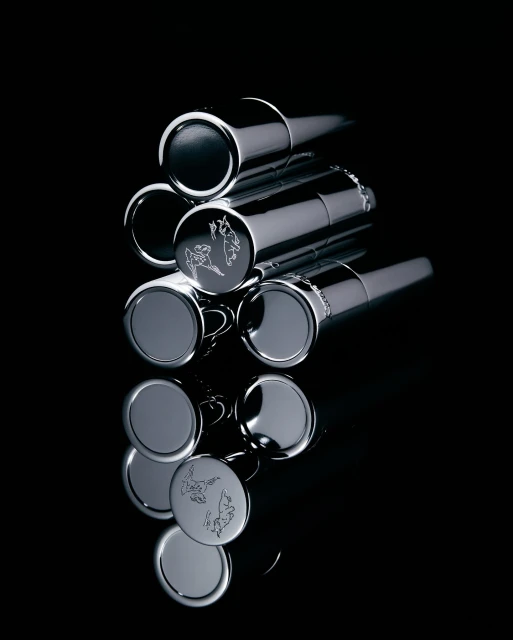 Stack of chrome metal lipstick cases or similar cosmetic containers on a black reflective surface, product photography, dark studio lighting.
