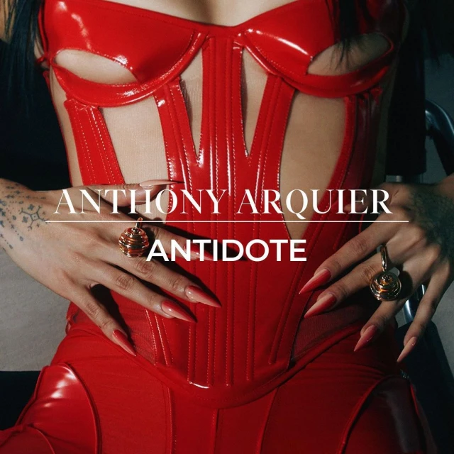 A woman in a red patent leather corset and dress poses with manicured hands, displaying large gold rings, against a dark background.
