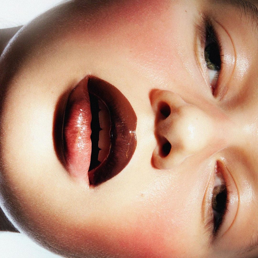 Close-up portrait of a young adult female face with open mouth, glossy lips, showing teeth, illuminated by soft lighting in studio.