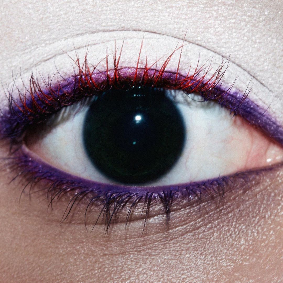 Close-up of a human eye with red and black eyelashes, purple eyeliner, and light skin, capturing beauty and cosmetic details.
