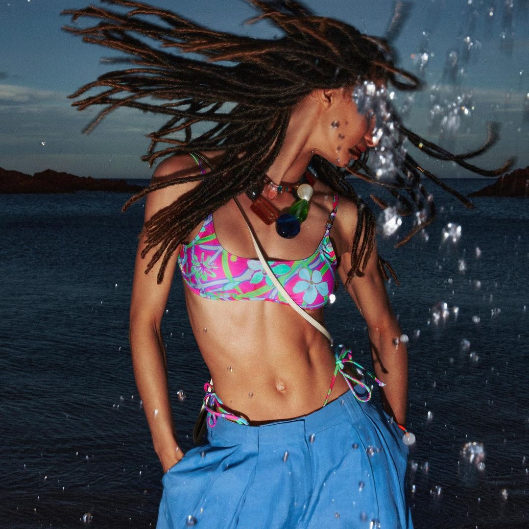 Woman with dreadlocks in bikini and blue pants near ocean with rocks, photographed with water droplets and a medium shot on a sunny day.