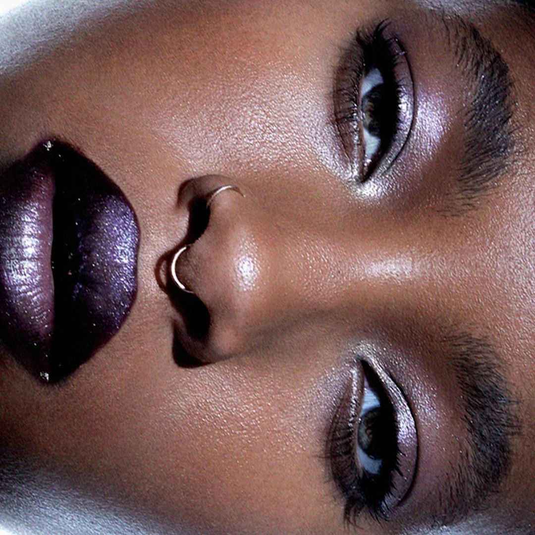 Close-up portrait of an adult female with brown skin, shiny lips, and nose piercings, emphasizing skincare and beauty details.