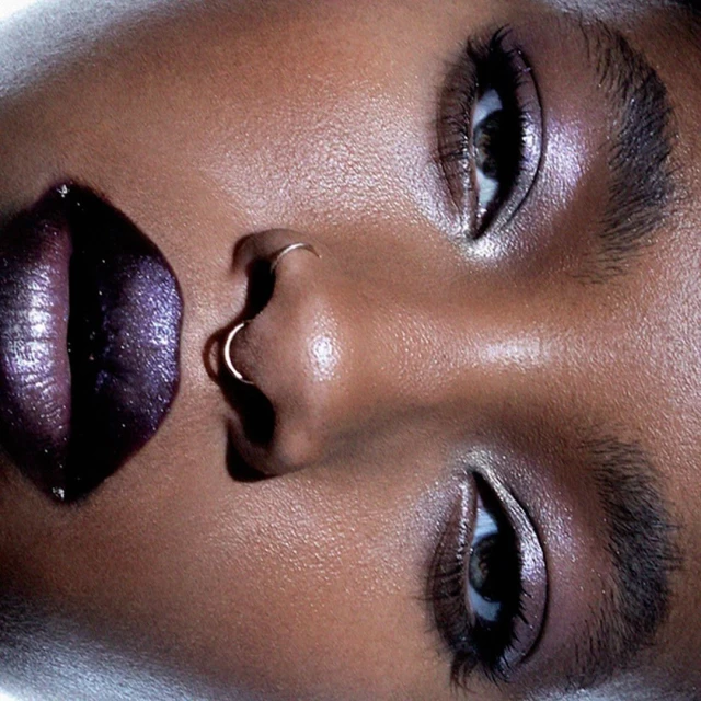 Close-up portrait of an adult female with brown skin, shiny lips, and nose piercings, emphasizing skincare and beauty details.